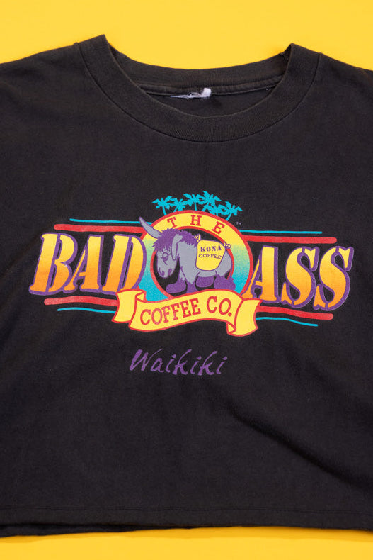 A black short-sleeve graphic t-shirt laid flat on a bright yellow background. The shirt features a colorful retro-style chest print for 'The Badass Coffee Co' with a rhinoceros logo and the word 'Waikiki' written below in purple script.