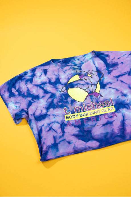 A short-sleeve tie-dye t-shirt in purple and blue, featuring a graphic of a bodybuilder with a barbell on a yellow moon and the text 't. micheal BODY BUILDING GEAR AMERICA', laid flat on a solid yellow background.