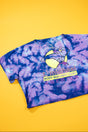 A short-sleeve tie-dye t-shirt in purple and blue, featuring a graphic of a bodybuilder with a barbell on a yellow moon and the text 't. micheal BODY BUILDING GEAR AMERICA', laid flat on a solid yellow background.
