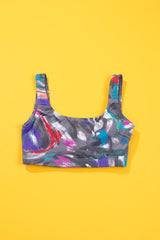 A sports bra with a colorful abstract paint-splatter pattern in purple, blue, pink, and white on a dark gray base, displayed flat against a solid yellow background.