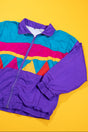 A vintage 1980s colorblock windbreaker jacket with purple sleeves and collar, teal upper body, and horizontal pink, yellow, and red stripes, laid flat on a bright yellow background.