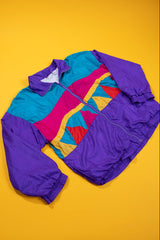A multicolor 1980s windbreaker jacket with purple sleeves and hem, a turquoise upper chest, and a geometric triangle pattern in pink, yellow, and red across the center, laid flat on a solid yellow background.
