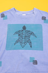 A light blue short-sleeve t-shirt featuring a central turquoise graphic of a black-and-white patterned figure inside a turquoise square, with small turquoise and black geometric accents on the sleeves and lower front, laid flat against a bright yellow background.