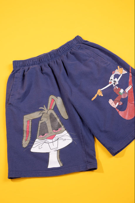 A pair of navy blue shorts featuring a large graphic of Bugs Bunny on the front and a smaller graphic of Looney Tunes characters on the side, displayed flat against a solid yellow background.