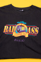A black short-sleeve graphic t-shirt laid flat on a bright yellow background. The shirt features a colorful retro-style chest print for 'The Badass Coffee Co' with a rhinoceros logo and the word 'Waikiki' written below in purple script.