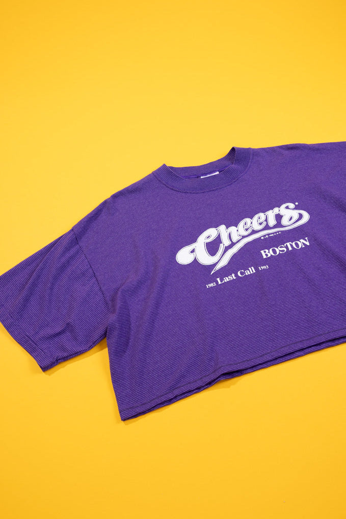 A purple cropped t-shirt with a white graphic reading 'Cheers Boston' and 'The Last Call 1991' laid flat on a solid yellow background.